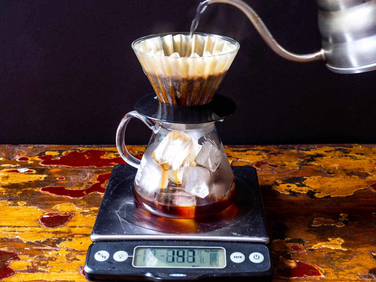 A kettle pouring hot water over coffee grounds in a cone filter dripping into a pitcher containing ice on a kitchen scale. 