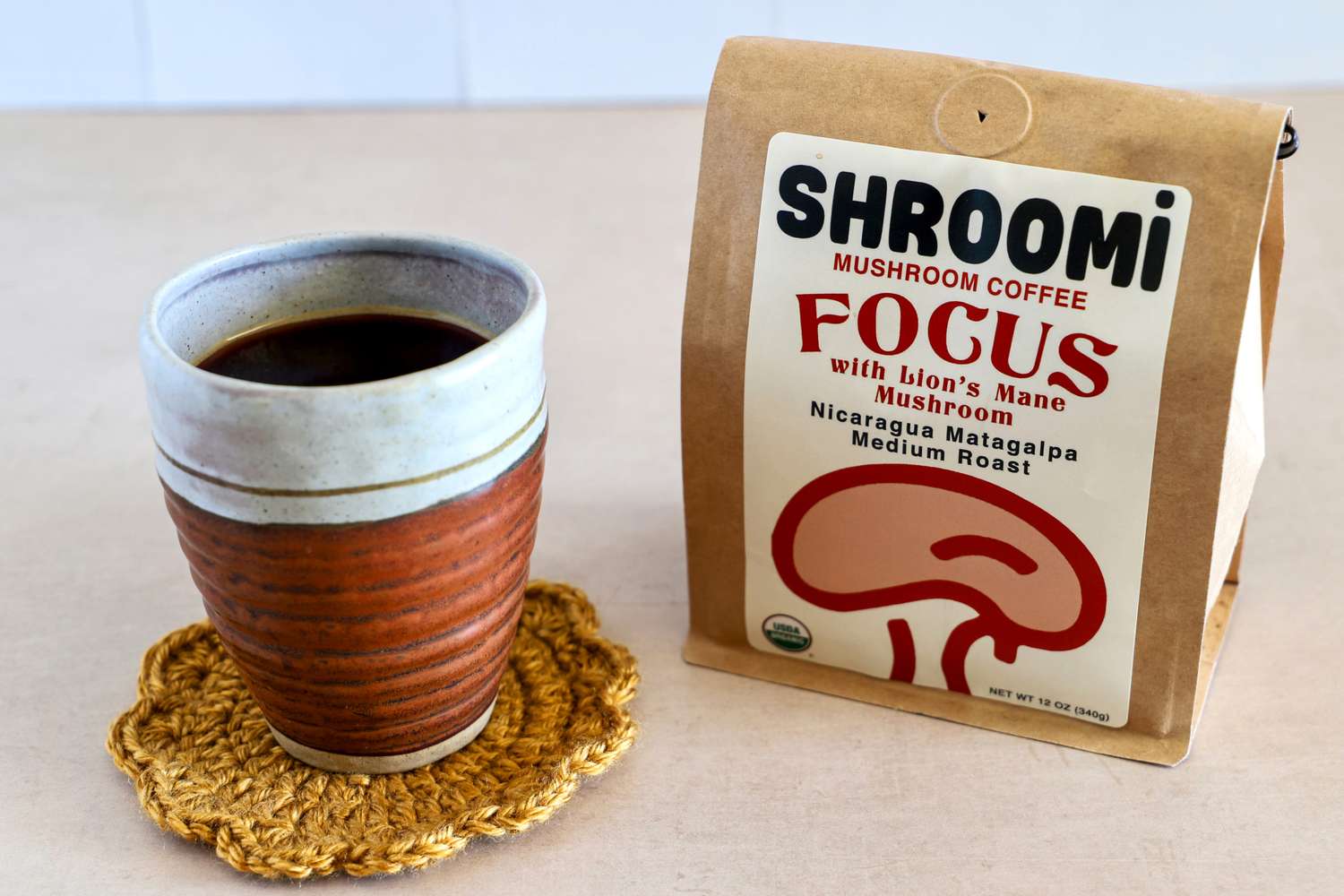 A ceramic cup filled with coffee on a crocheted coaster next to a bag labeled Shroomi Mushroom Coffee Focus with Lion's Mane