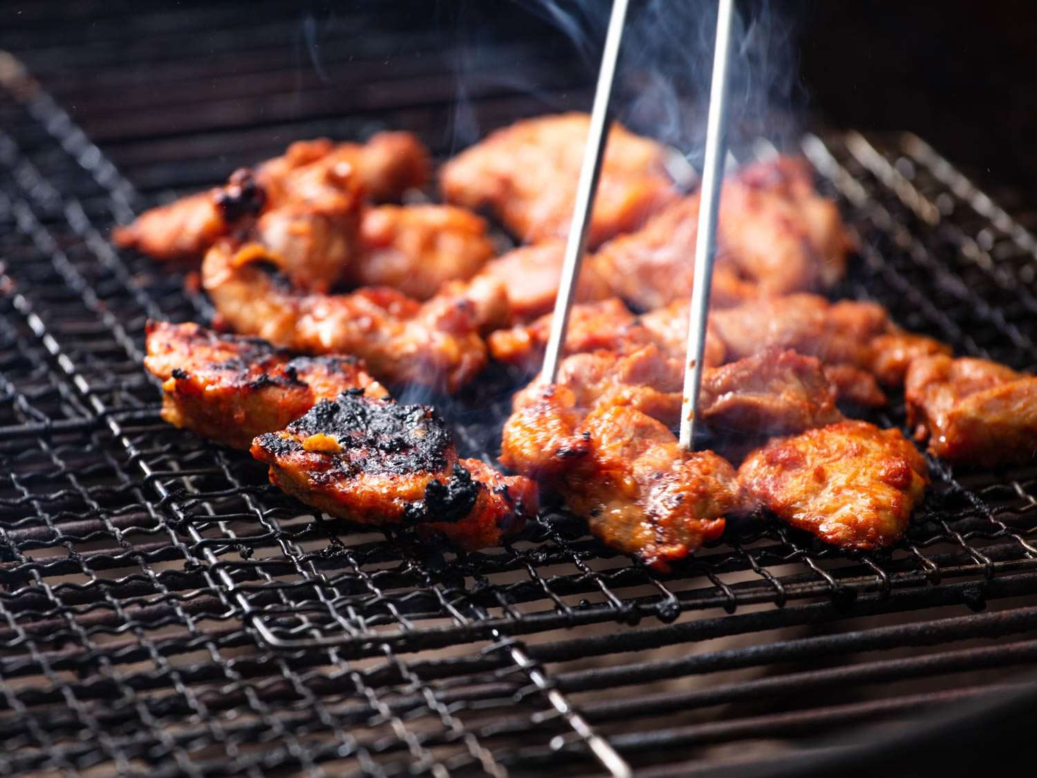 Turning chunks of pork shoulder on the grill with kitchen tweezers.