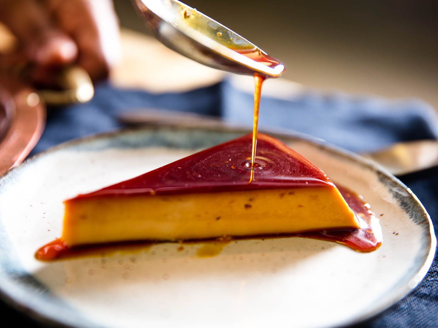 A spoon drizzling caramel sauce onto a wedge of flan on a serving dish. 