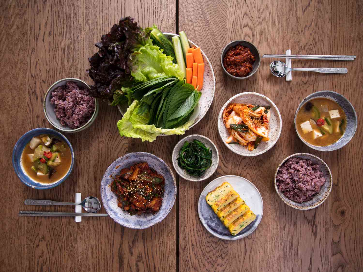 Overhead view of a wooden table full of Korean banchan, purple sticky rice, bowls of soup, and an assortment of lettuce leaves, perilla leaves, carrot sticks, and lengths of quartered cucumber.