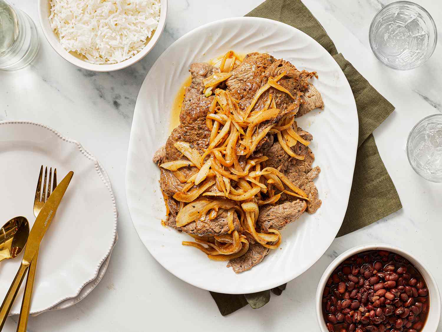 A plated meal featuring bistec encebollado with onion garnish, served with rice and red beans on the side