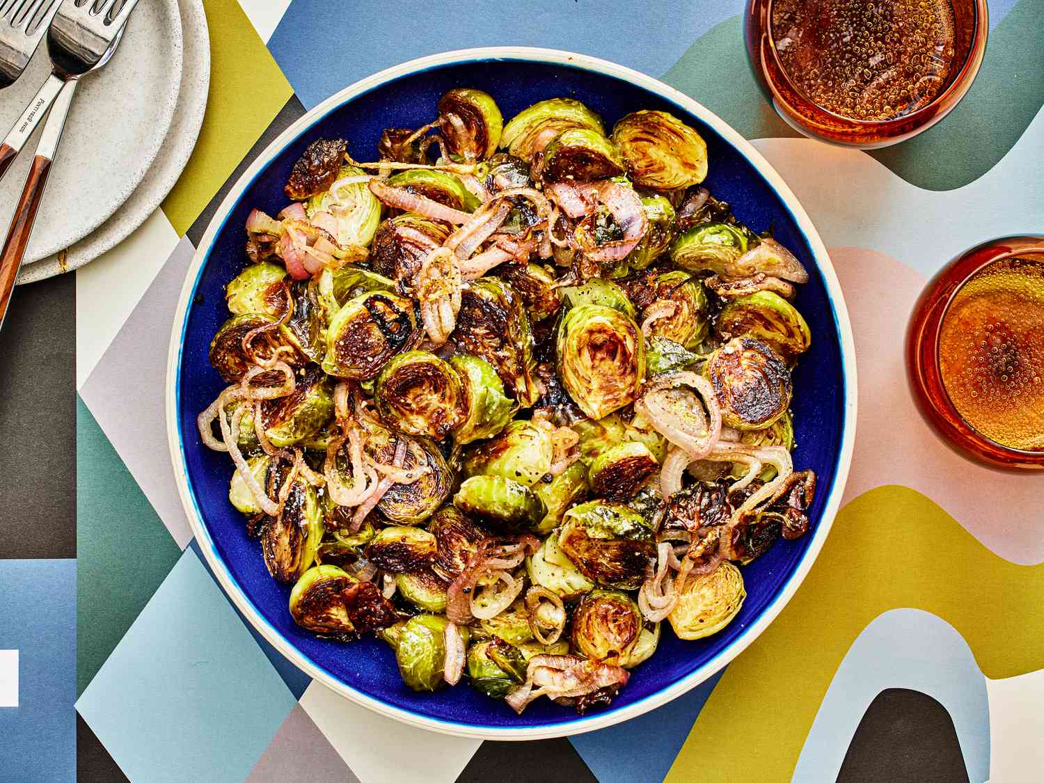 Roasted brussel sprouts and shallots with a balsamic glaze in a bowl, with glasses of sparkling water, some smaller plates and a colorfully printed table top