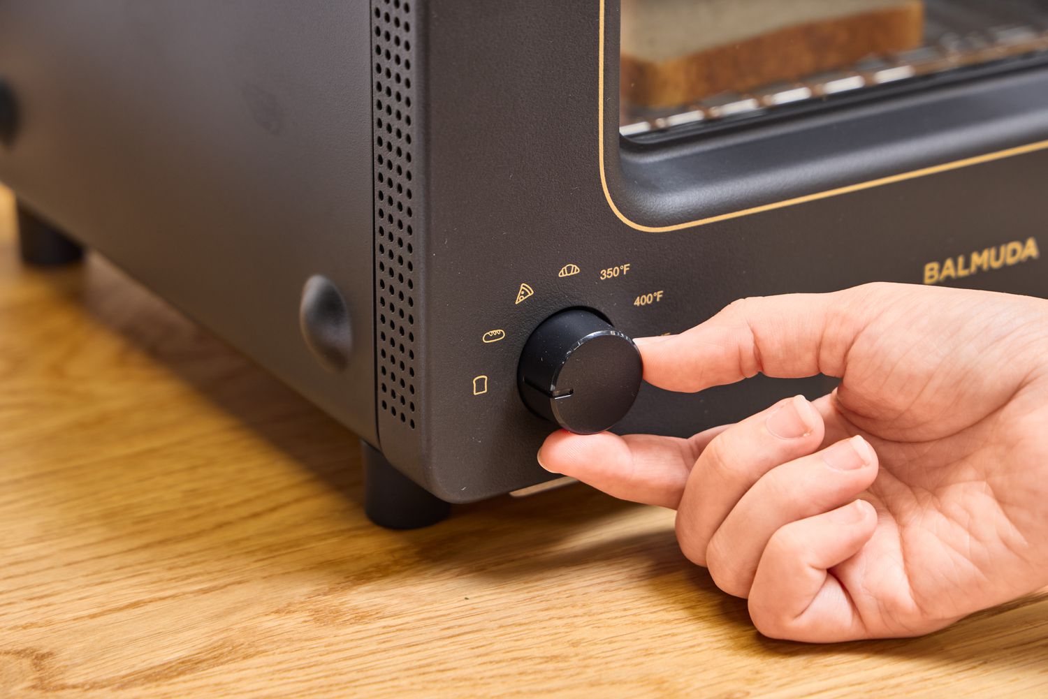 A hand turning a temperature dial on a balmuda toaster oven with symbols for different food settings