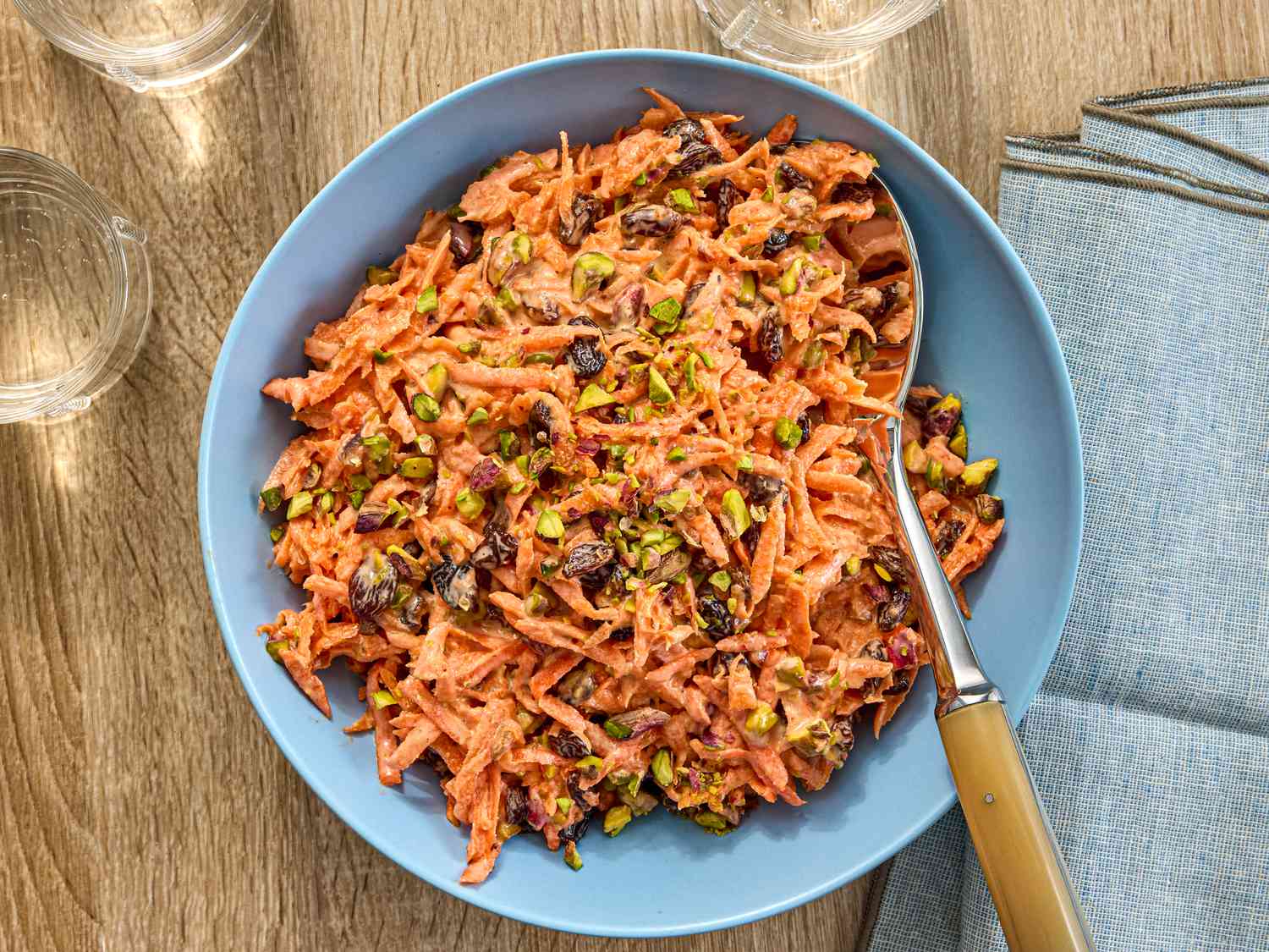 Carrot slaw in a blue bowl surrounded by glasses of water on a wooden surface with napkins and serving spoon 