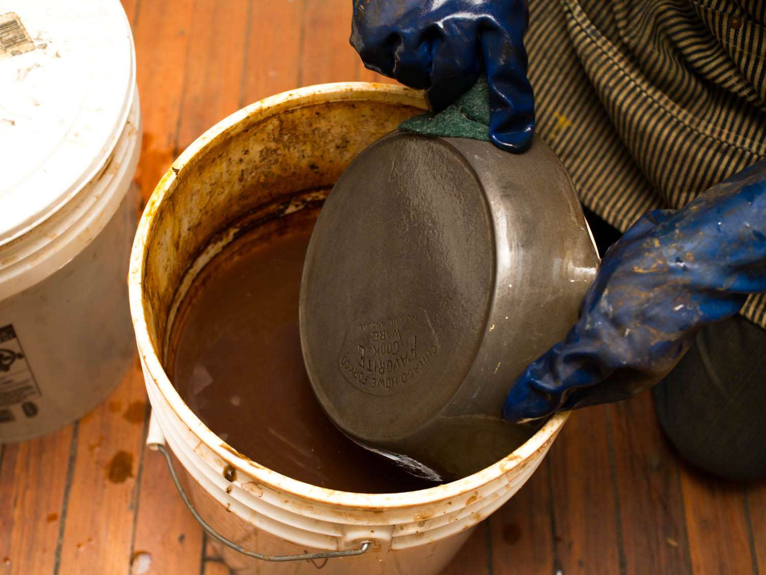 Gloved hands pulling a cast iron skillet from a bucket of soaking solution. 
