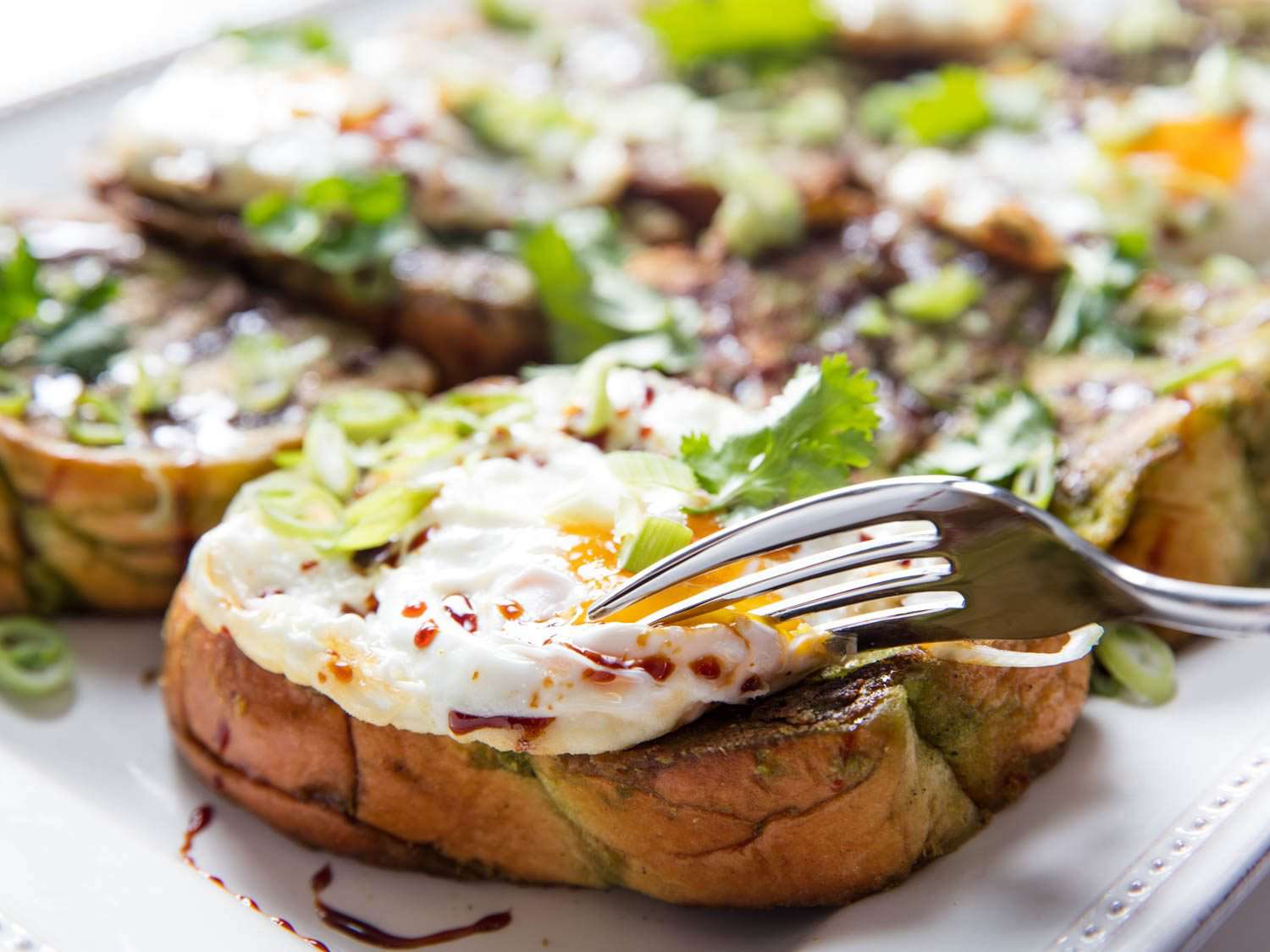 Close-up of a fork cutting into a fried egg on top of savory green curry French toast, with more French toast in the background