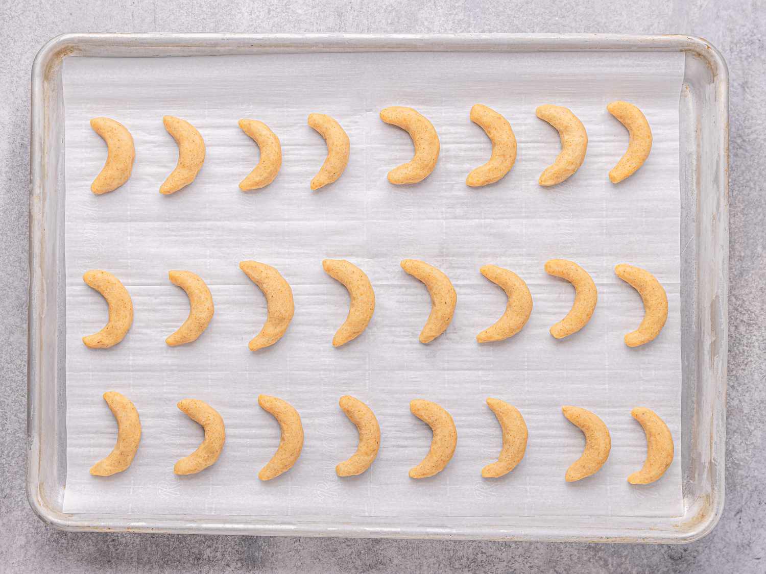 Baking tray arranged with crescentshaped cookies in rows on parchment paper