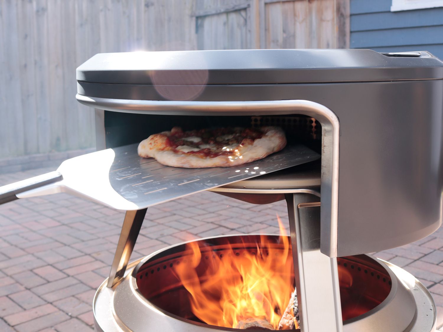 A pizza being loaded into an outdoor pizza oven on a metal peel 