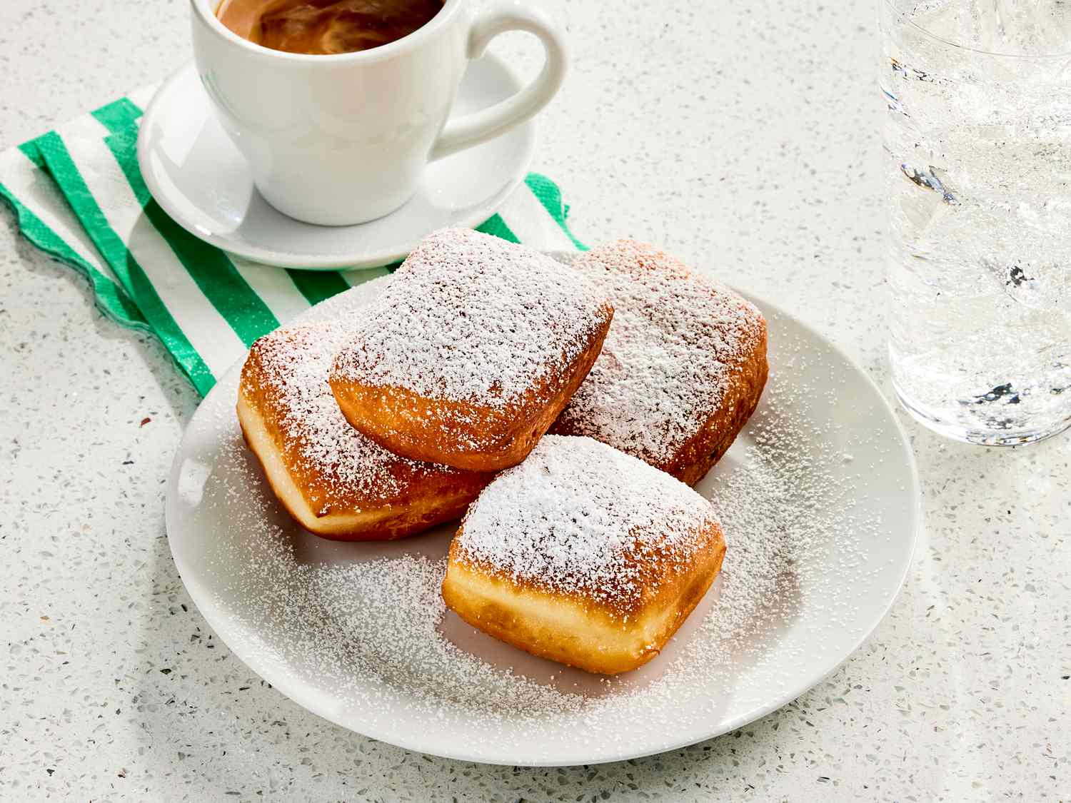 Beignets on a white plate with powdered sugar, with a coffee, green and white striped napkin, and a marble surface