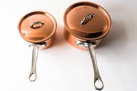 Two copper saucepans with lids featuring silver handles placed side by side on a white surface