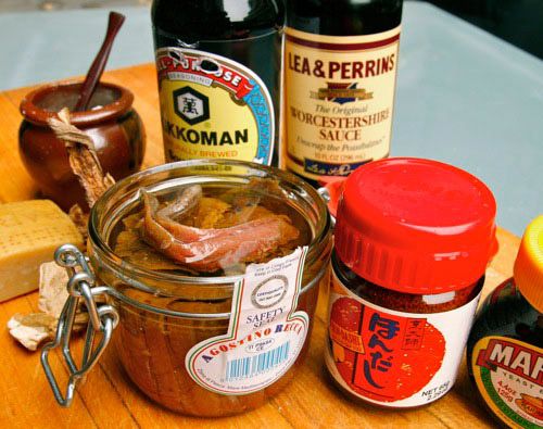 Jars of anchovies, Hondashi, marmite, MSG, soy sauce, and Worcestershire sauce alongside a hunk of parmesan cheese.