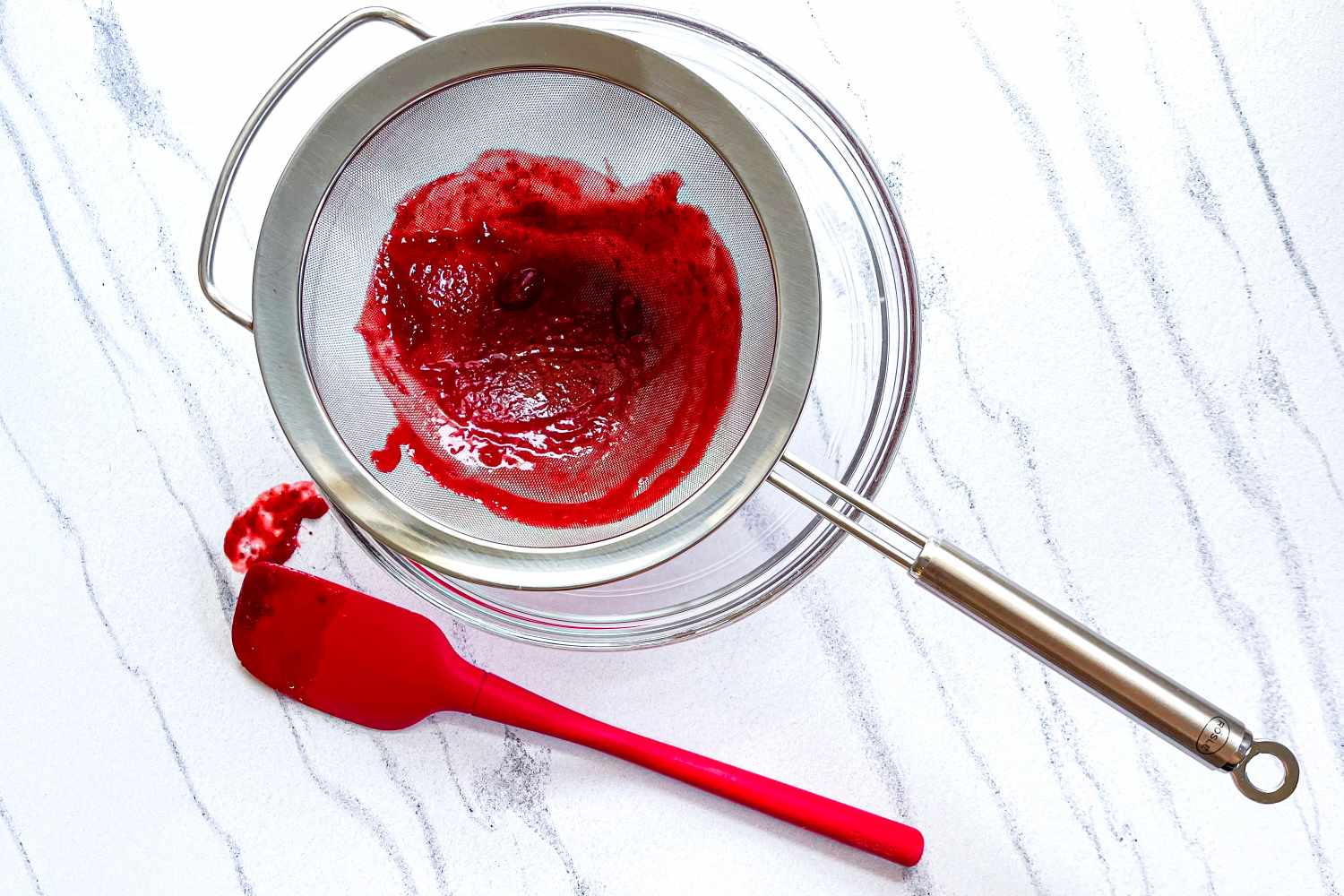 A red spatula beside the Rösle Stainless Steel Round Handle Kitchen Strainer with red puree on a clean surface