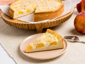 A slice of peach streusel cake on a plate with a larger cake in the background on a wooden stand
