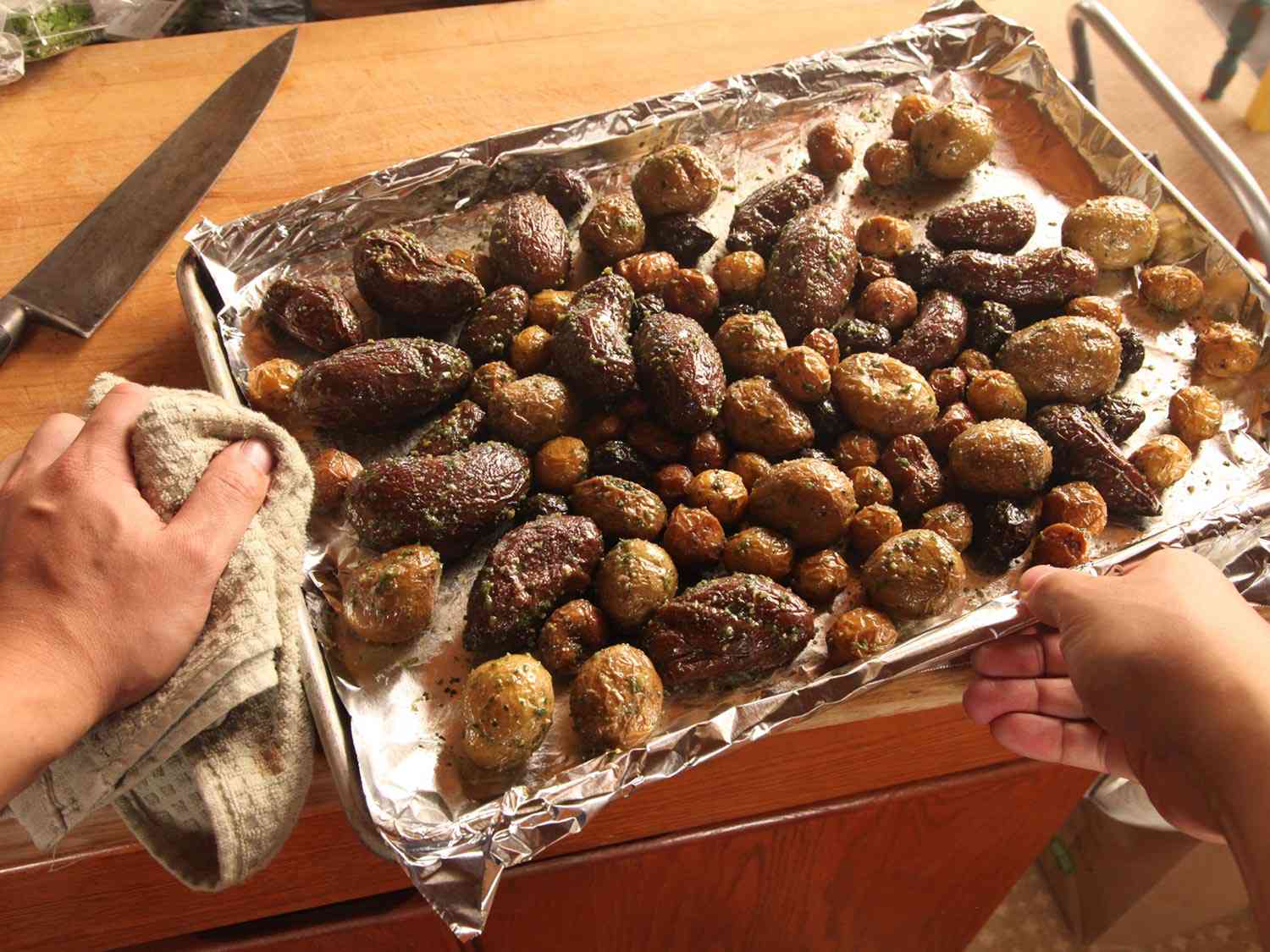 Two hands holding a sheet pan filled with roasted potatoes of various sizes.