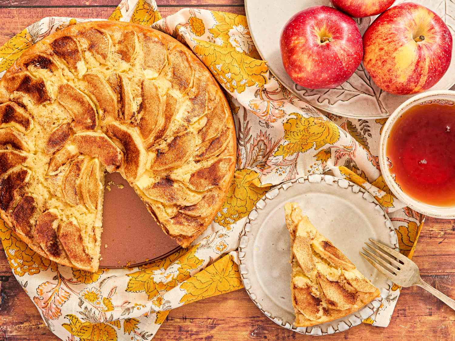 A sliced apple cinnamon cake on a decorated table with apples and tea nearby