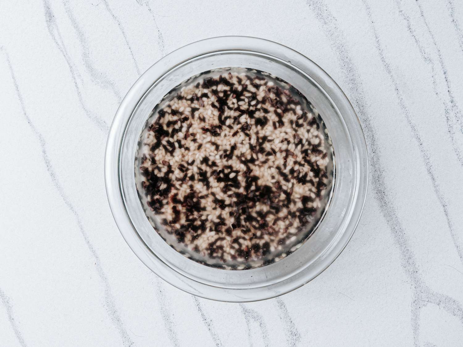 Rice sitting in a glass bowl of water on a white marbled surface 