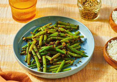 A plate of green beans seasoned with spices, next to a bowl of rice and a jar of seasonings on a textured surface