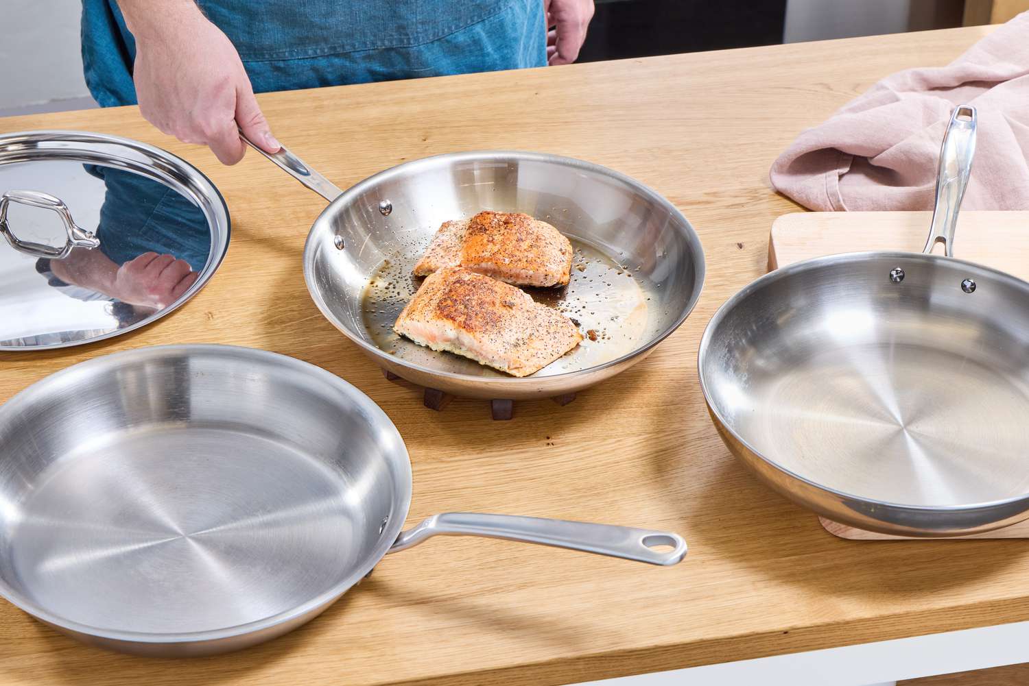 Three stainless steel skillets on a kitchen countertop. One has two cooked salmon fillets in it.