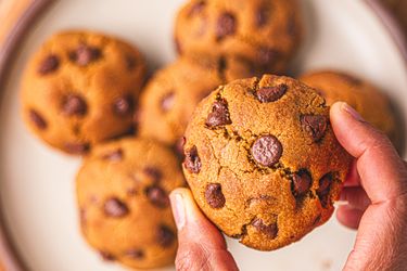 A closeup of hands holding a chocolate chip cookie with other cookies visible on a plate in the background
