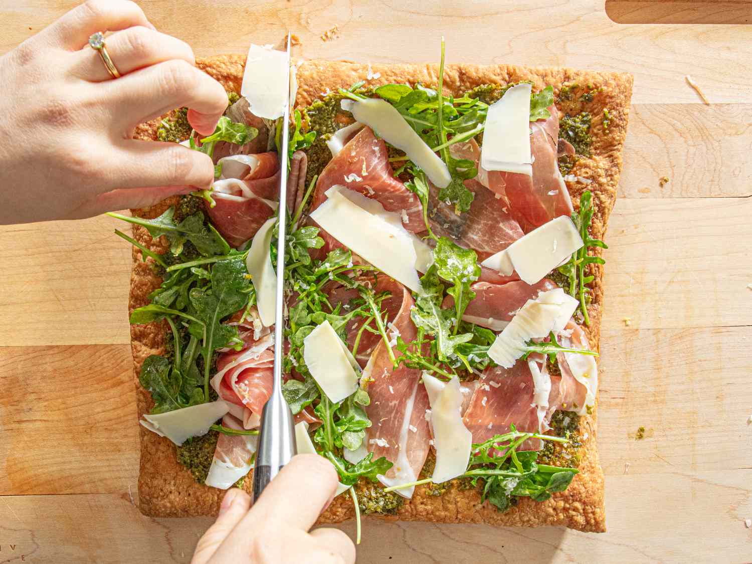 A charcuterie flatbread being cut into slices topped with prosciutto arugula and shaved cheese