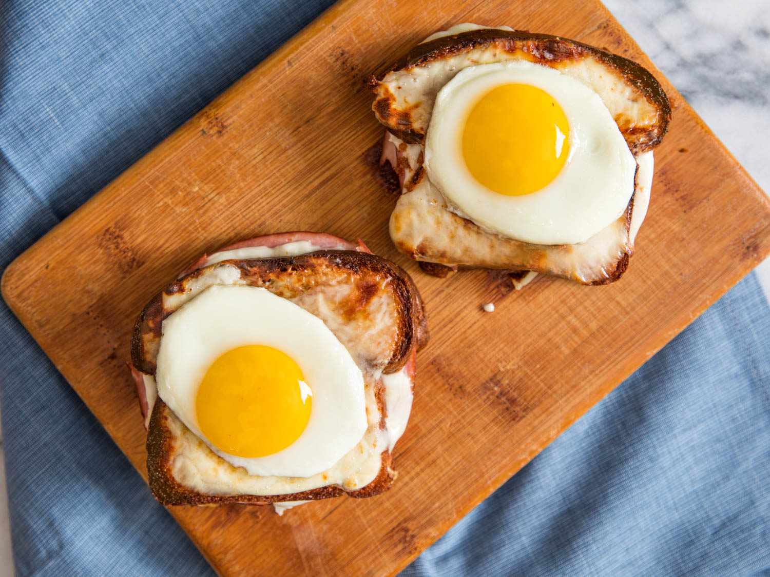 Two Croque Madame sandwiches, topped with sunny-side up eggs on a wooden board on a blue cloth.