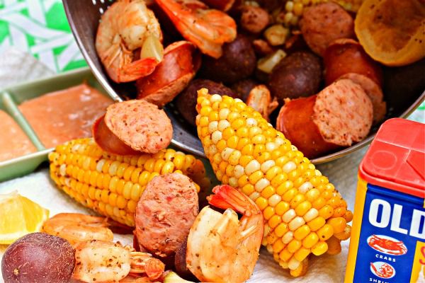 Closeup of a colander full of low-country shrimp boil on its side, cornucopia-like, with shrimp, halved corn cobs, and chunks of sausage spilling forth. Old Bay seasoning and dipping sauce are visible on either side.