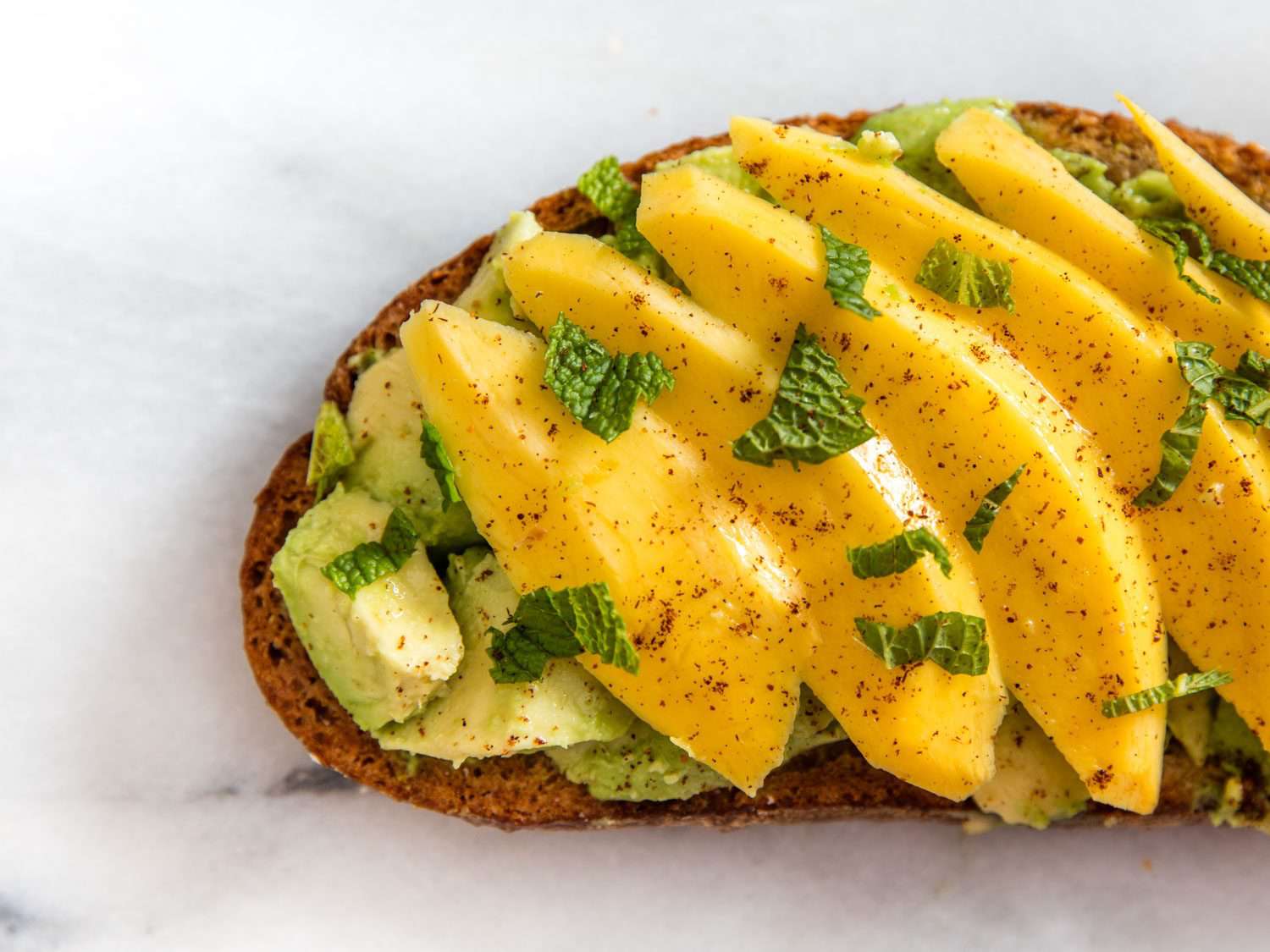 Close-up of avocado toast topped with sliced mango, mint, and chile powder.