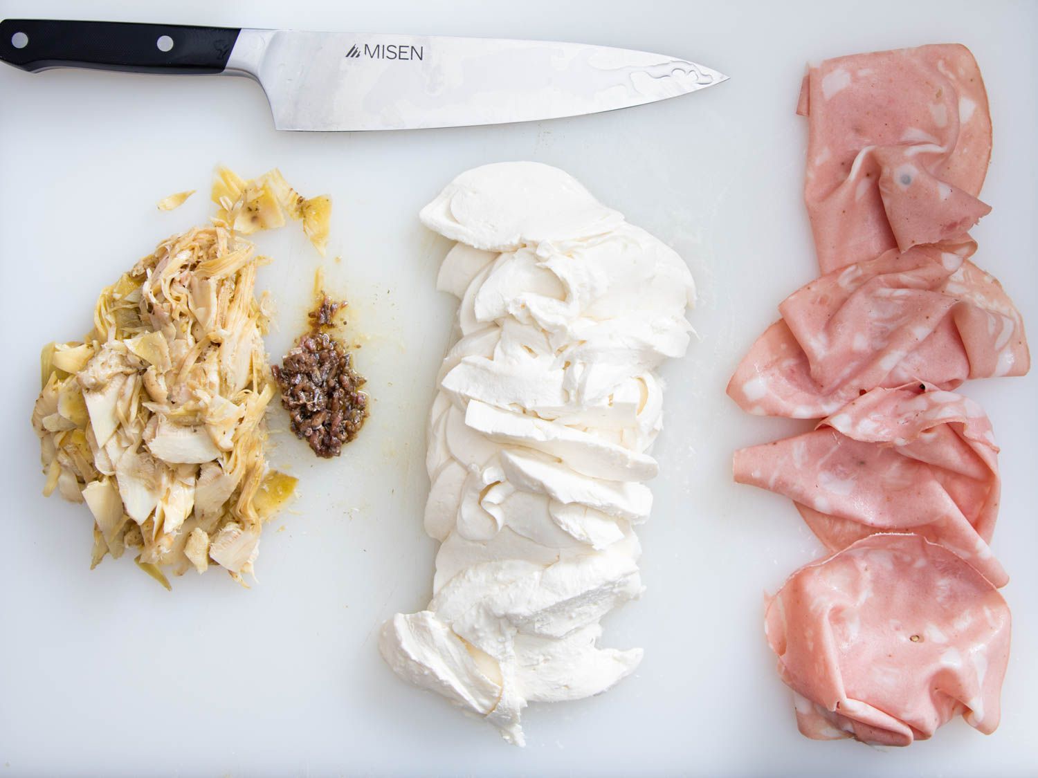Overhead view of the ingredients for the sandwich arranged in rows on a white surface: chopped marinated artichoke hearts, fresh mozzarella, thin slices of mortadella, and a small mound of minced anchovy.