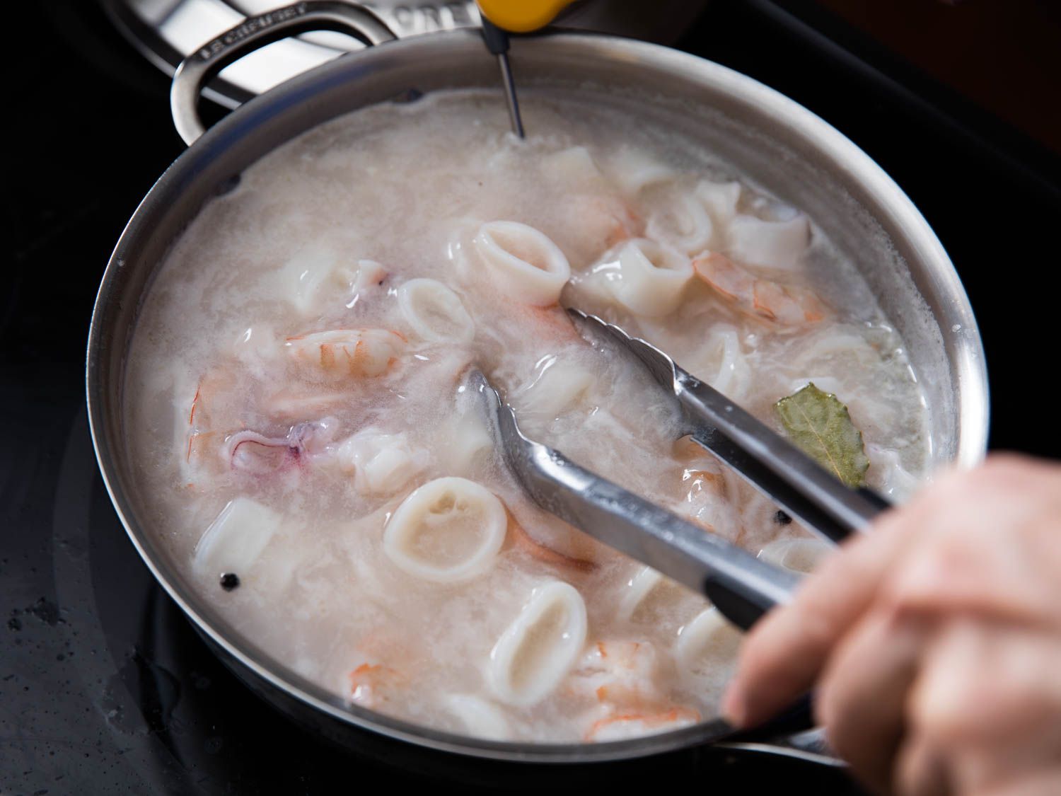 Poaching squid and shrimp in a skillet being stirred with tongs.