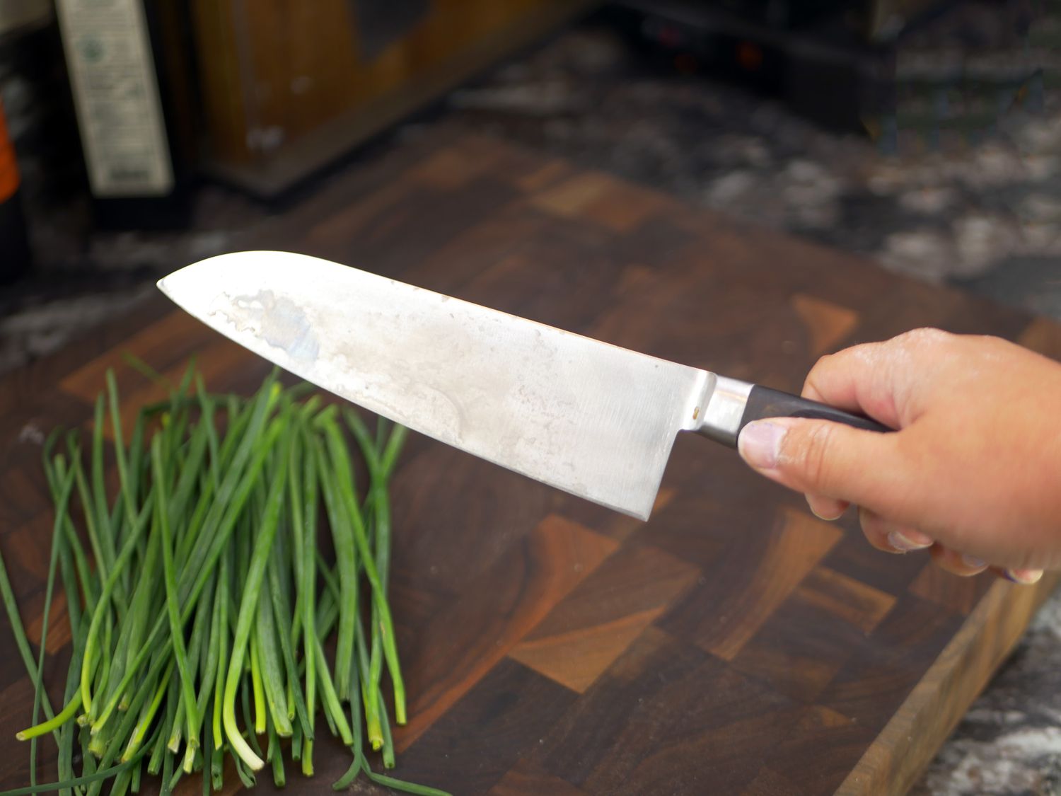 a person holding a santoku knife over a cutting board with chives on it 