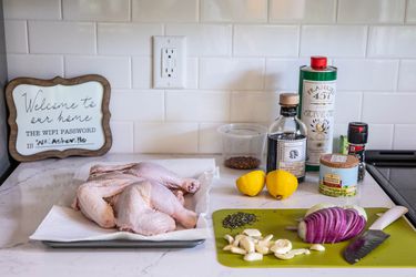 A spread of ingredients on a marble countertop in an Airbnb kitchen: chicken parts on a paper towel–lined tray; onion, garlic, and herbs on a cutting board; balsamic vinegar; fleur de sel; olive oil; grinder of black pepper; lemon halves