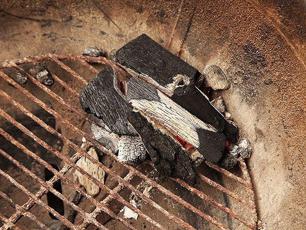 A small mound of half-lit lump charcoal placed along the wall of a kettle-style grill. Some coals on the bottom are glowing and the topmost pieces are beginning to catch.