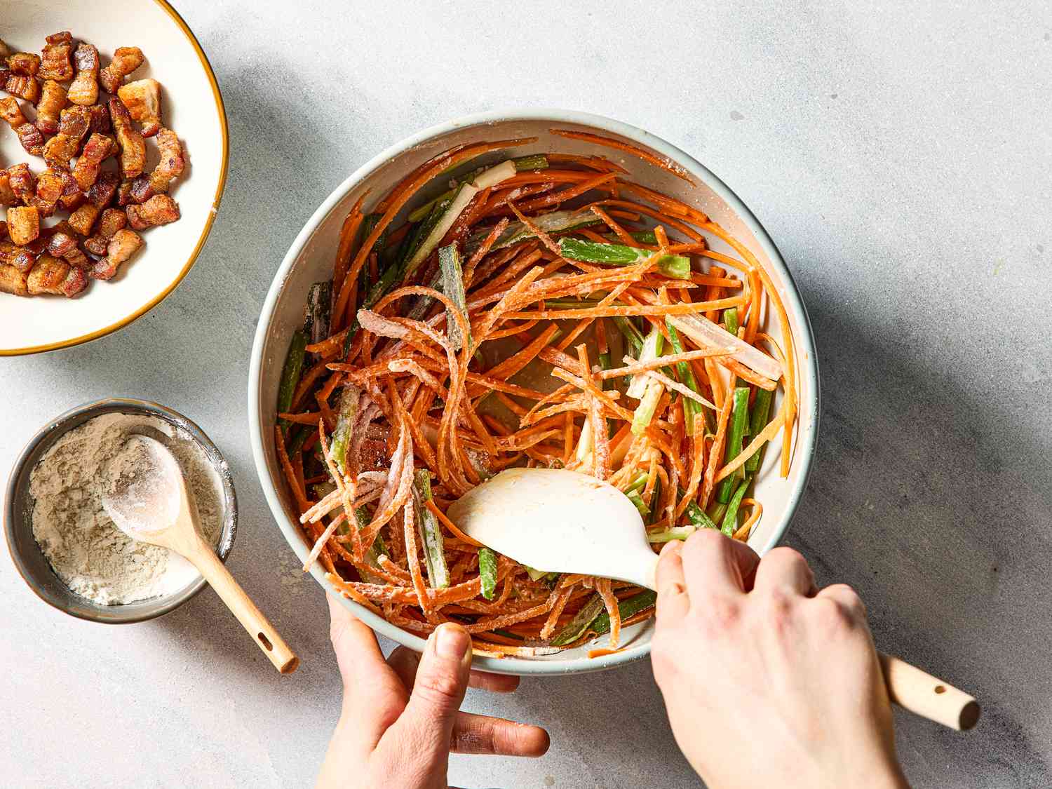 A bowl of mixed ingredients including shredded vegetables being stirred, a plate of diced meat and a small bowl of flour nearby