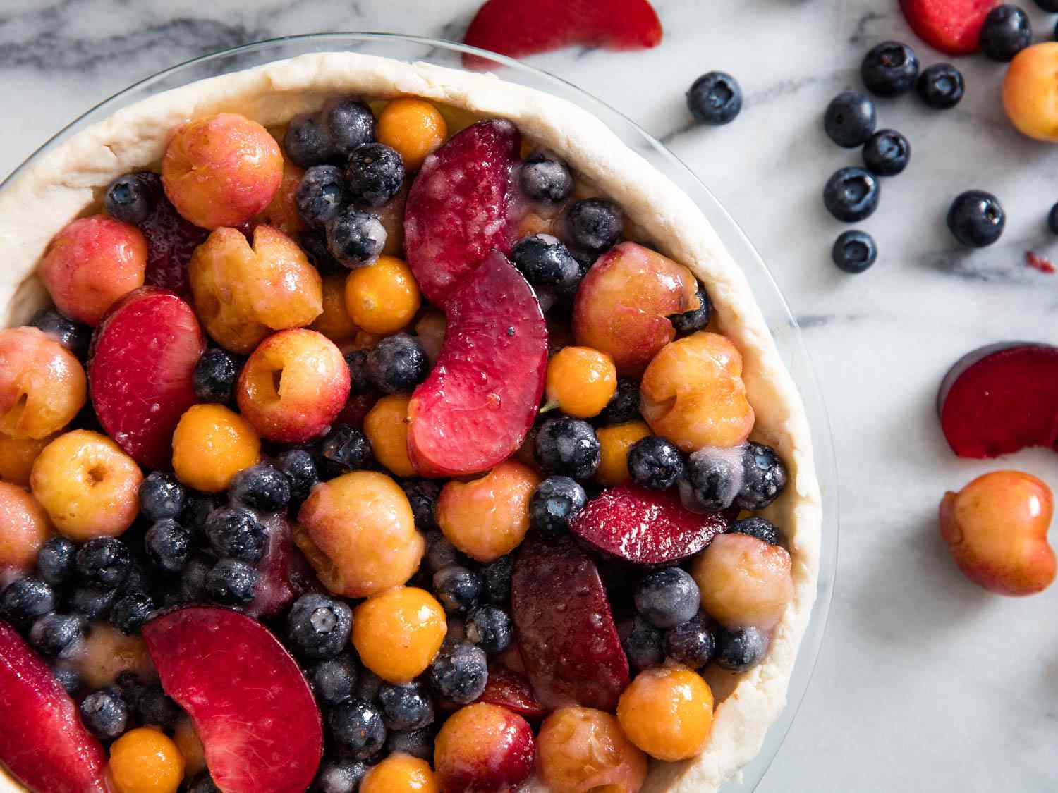 Overhead shot of a pie dish on a white marble slab. The pie dish is lined with dough and filled with blueberries, Cape gooseberries, Rainier cherries, and plum slices. The fruit has been tossed into the sugar-starch mixture and extra fruit is scattered to one side of the dish.