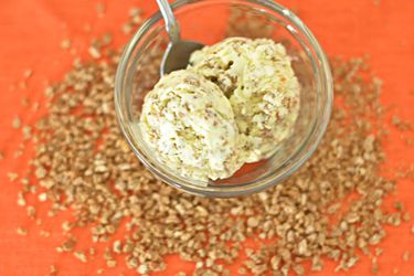 Overhead view of two scoops of Grape-Nuts ice cream served in a small bowl. Grape-Nuts are scattered on the orange surface below.