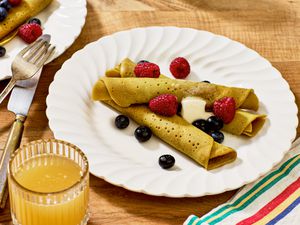 ployes with butter and berries on white dishes, with a fork and knife, striped napkin, and a wooden tabletop