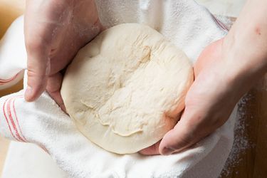 Two hands are placing a ball of bread dough into a bowl lined with a floured kitchen towel. 