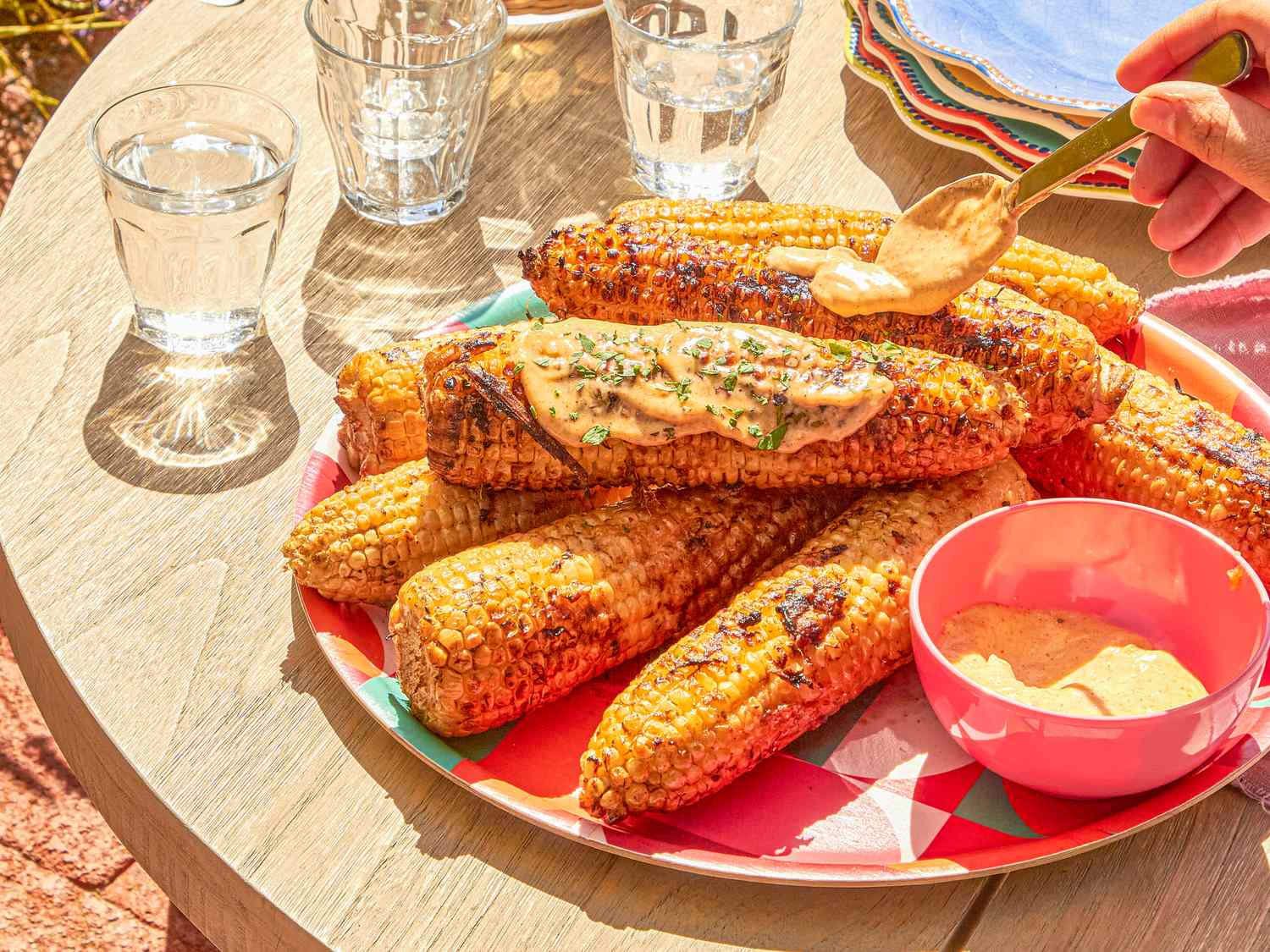 Grilled corn on a plate with sauce being applied table setting with water glasses and a bowl of sauce