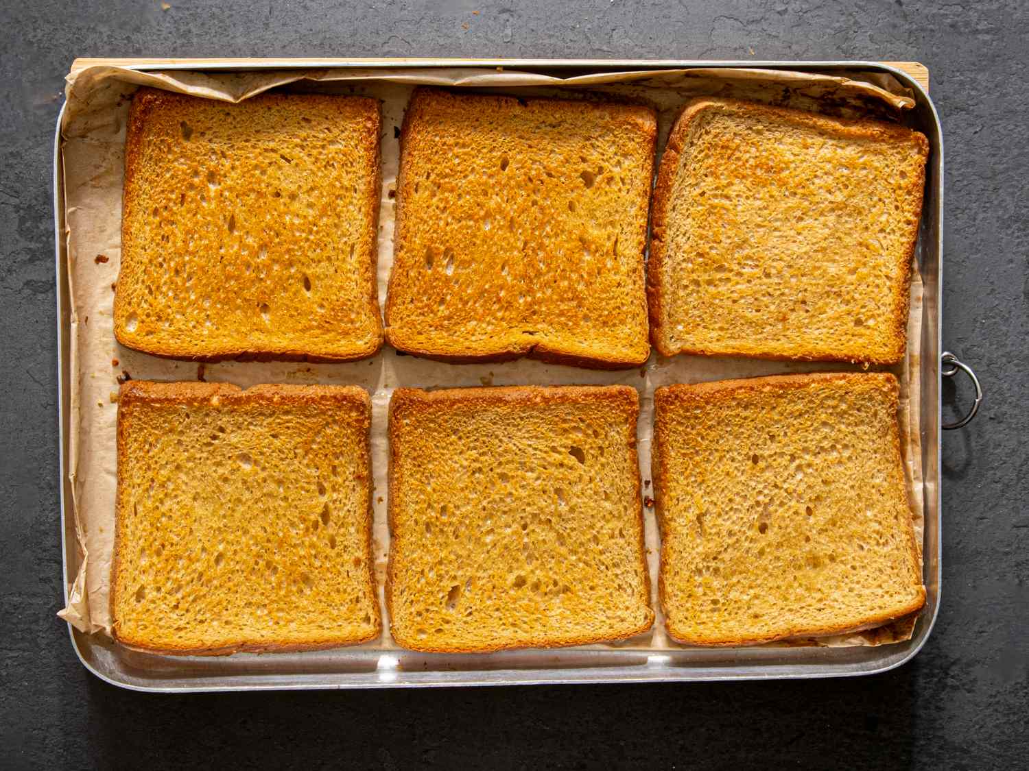 Six slices of toasted bread arranged on a baking tray lined with parchment paper