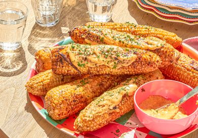 Plated grilled corn cobs topped with sauce and herbs alongside a small bowl of sauce