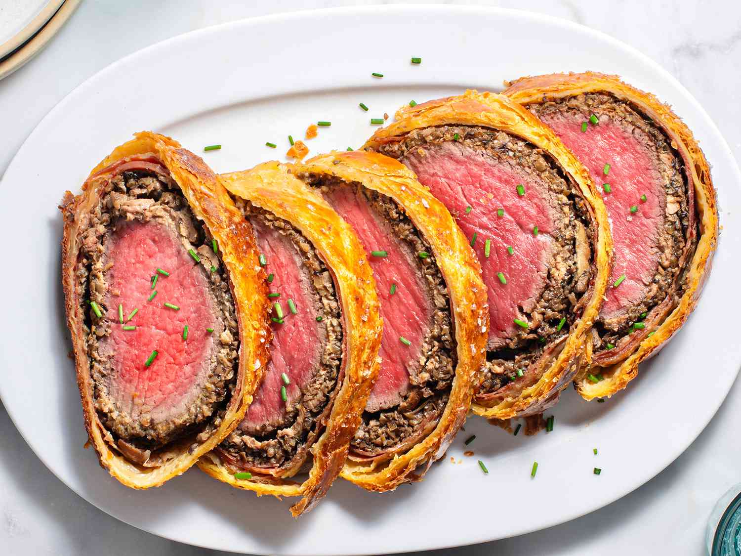 A white oval ceramic plate holding five slices of Beef Wellington which are sprinkled with chopped chives.