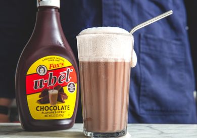A glass of chocolate egg cream with a metal straw next to a bottle of Ubet chocolate syrup
