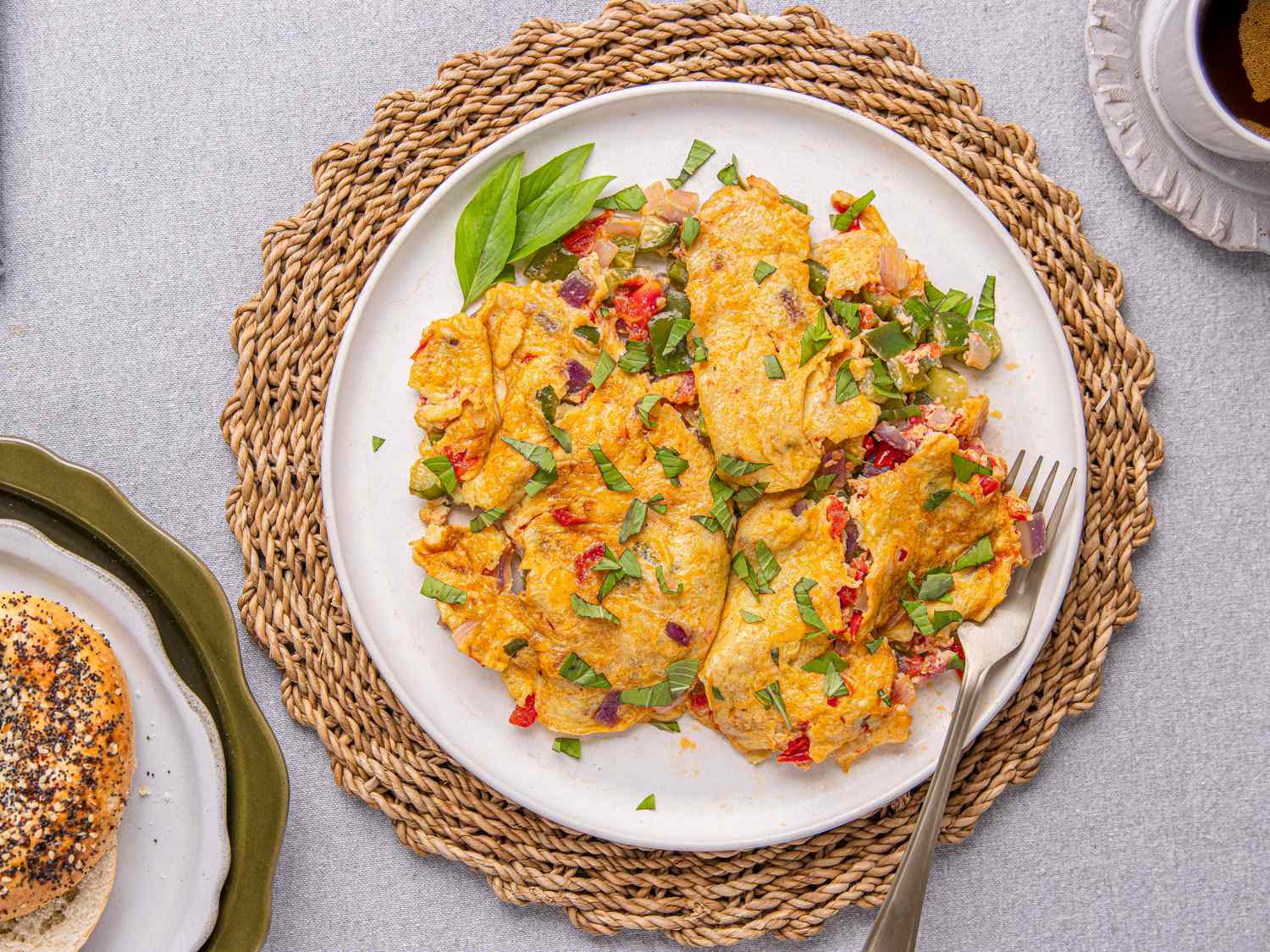 A plate of a prepared Nigerianstyle omelet garnished with herbs accompanied by a bagel and a cup of tea on a woven mat