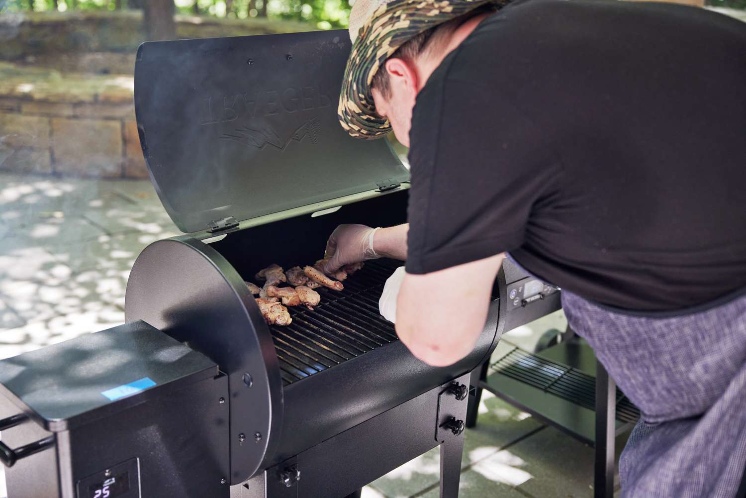 a person hunched over and adding chicken wings to a smaller pellet grill
