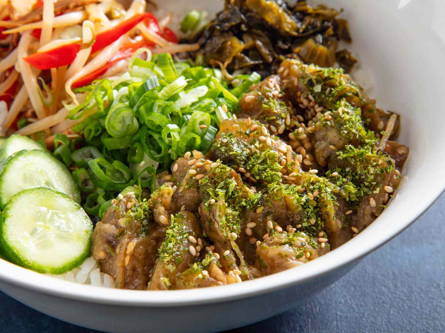A close-up of a rice bowl topped with braised eggplant, cucumbers, chopped scallions, and bean sprouts.