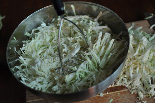 Salted cabbage shreds are lightly pounded with a potato masher in a large mixing bowl.