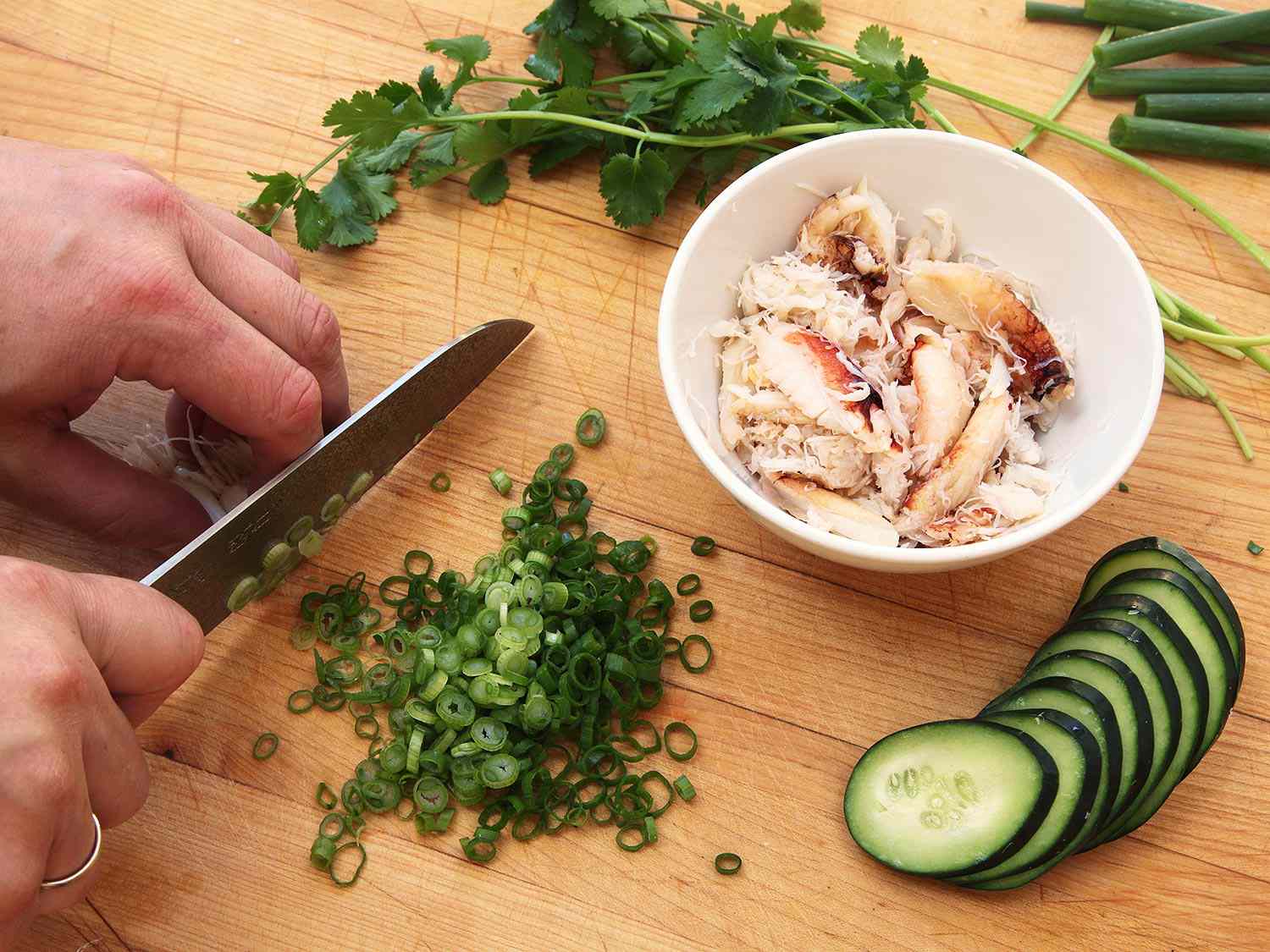 The ingredients for crab fried rice are prepped on a cutting board: scallions are being sliced by the author; picked crabmeat, sliced cucumber, and cilantro are nearby.