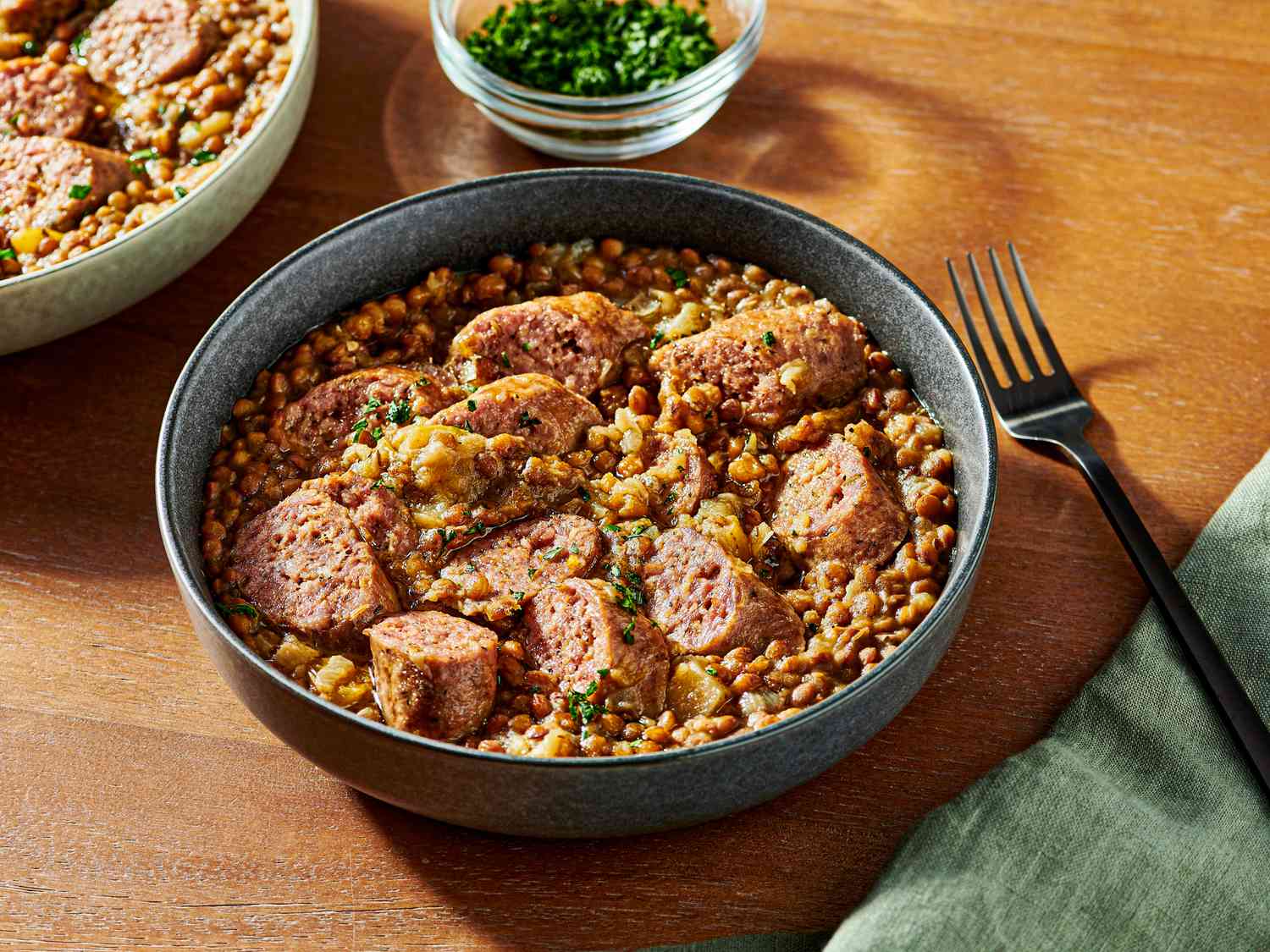 2 bowls of pressure cooked lentils and sausage on a wooden tabletop, with a soft turquoise napkin, a fork, and a small bowl of parsley for garnish to the sides 