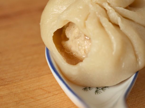 Close-up of a finished xiao long bao dumpling on a soup spoon. A small bite has revealed the soupy interior and ball of filling swimming inside.